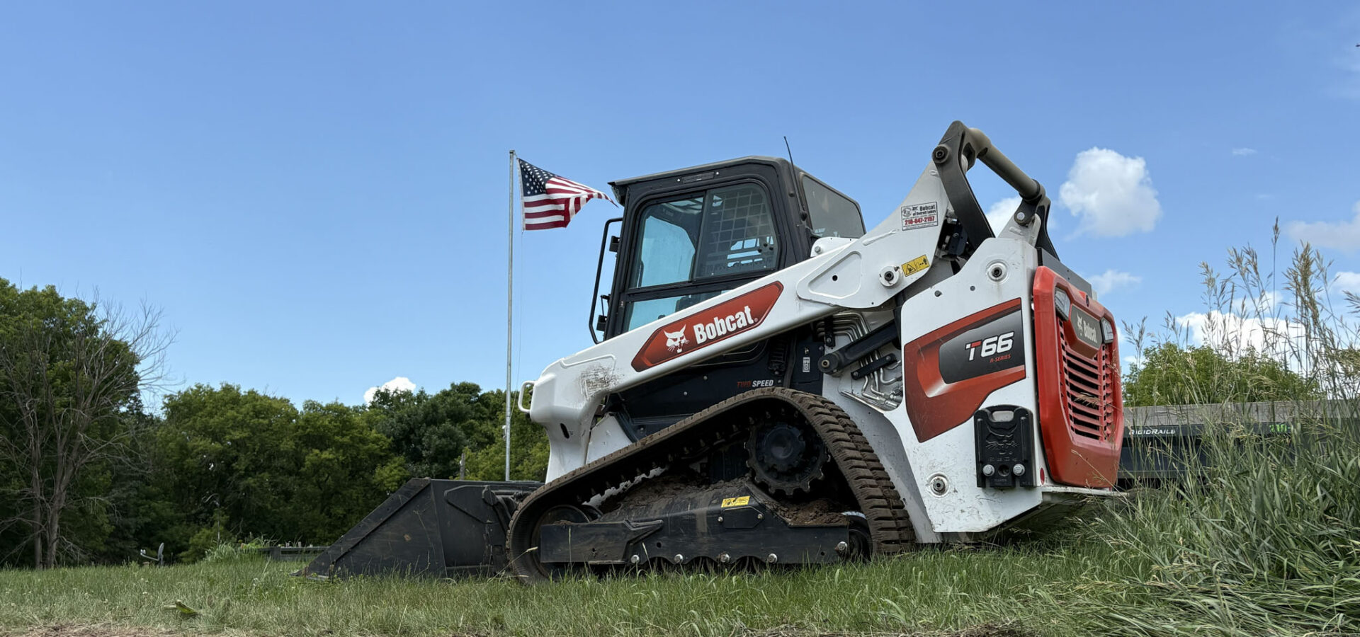 : Bobcat T66 compact track loader parked on grassy terrain with an American flag in the background under a blue sky.